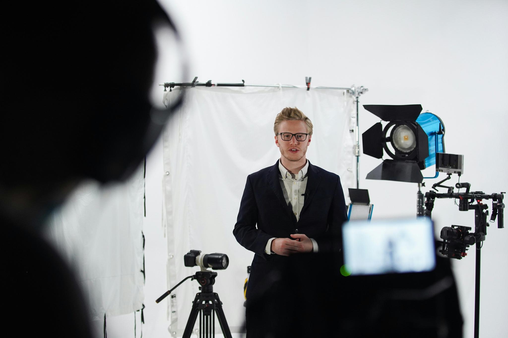 Un homme en costume avec des lunettes dans un lieu de tournage face à la caméra